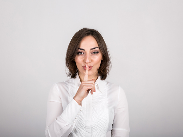 A imagem mostra uma mulher branca de olhos claros e cabelos castanhos com o dedo indicados sobre a boca em uma indicação de silêncio. Ela veste uma camisa branca de botões. O fundo da imagem é uma parede branca.