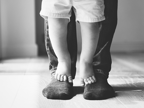Imagem em preto e branco de um bebê em pé caminhando sobre os pés do pai. O fundo da foto é desfocado.