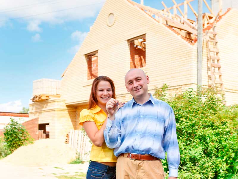 Casal feliz em frente a uma casa em processo de construção. A mulher é branca, tem cabelo ruivo e liso, e veste uma camisa amarela. O homem é branco, careca, e veste camisa azul e calça bege.