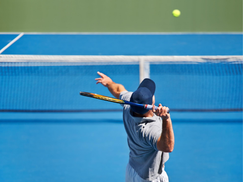 Homem de pele bronzeada, camiseta curta e bermuda, saltando com uma raquete de tênis na mão. Ao fundo, uma quadra de tênis em um dia ensolarado.
